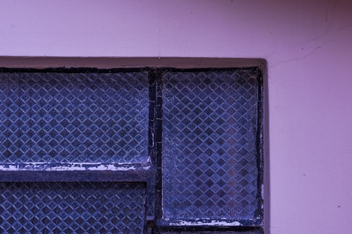 A textured, patterned glass window with a metal frame, surrounded by a light purple wall with visible cracks. The window glass displays a grid-like diamond pattern.