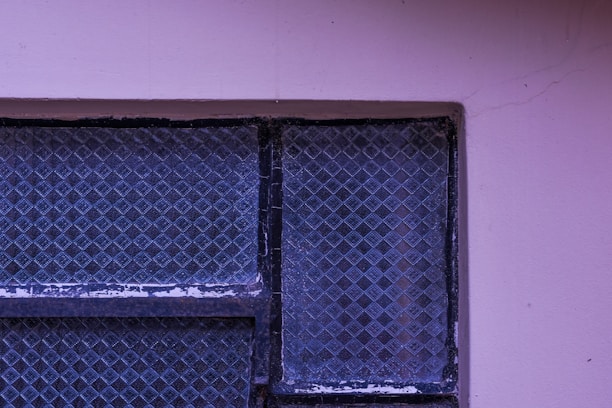 A textured, patterned glass window with a metal frame, surrounded by a light purple wall with visible cracks. The window glass displays a grid-like diamond pattern.