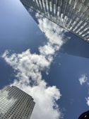 Tall modern skyscrapers with reflective glass panels towering against a blue sky. Fluffy white clouds are visible both in the sky and reflected on the surfaces of the buildings.