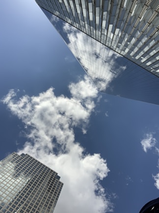 Tall modern skyscrapers with reflective glass panels towering against a blue sky. Fluffy white clouds are visible both in the sky and reflected on the surfaces of the buildings.