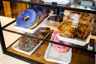 A display case showcases a variety of pastries. There are trays containing muffins and donuts with different toppings. The donuts feature bright glazes in blue, chocolate with sprinkles, and pink with sprinkles. The muffins are wrapped in brown paper.