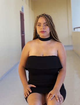 A person with braided hair, wearing a black off-shoulder dress with a choker and sitting in a corridor with beige walls and a maroon door in the background.