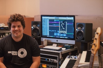 A person is sitting in a music studio with a setup consisting of a computer, studio monitors, and various audio equipment. The person is smiling and wearing a 'Record Time' t-shirt. The monitor displays music production software, and there are various musical instruments and devices around, including a keyboard and an amplifier.
