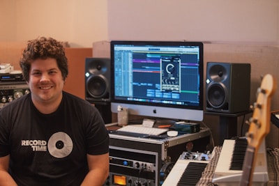 A person is sitting in a music studio with a setup consisting of a computer, studio monitors, and various audio equipment. The person is smiling and wearing a 'Record Time' t-shirt. The monitor displays music production software, and there are various musical instruments and devices around, including a keyboard and an amplifier.