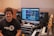 A person is sitting in a music studio with a setup consisting of a computer, studio monitors, and various audio equipment. The person is smiling and wearing a 'Record Time' t-shirt. The monitor displays music production software, and there are various musical instruments and devices around, including a keyboard and an amplifier.