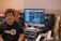 A person is sitting in a music studio with a setup consisting of a computer, studio monitors, and various audio equipment. The person is smiling and wearing a 'Record Time' t-shirt. The monitor displays music production software, and there are various musical instruments and devices around, including a keyboard and an amplifier.