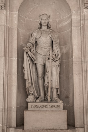 A stone statue of a crowned man in regal armor is set within an arched niche, detailed with a draped cloak and a sword held prominently. The base of the statue features the inscription 'Fernando El Católico'.