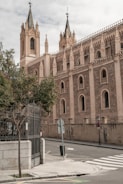 A Gothic-style church with ornate spires and detailed stone carvings, situated on a quiet street corner with a metal gate, trees, and a crosswalk.
