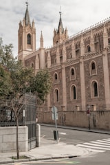 A Gothic-style church with ornate spires and detailed stone carvings, situated on a quiet street corner with a metal gate, trees, and a crosswalk.