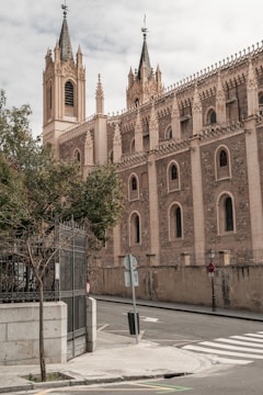 A Gothic-style church with ornate spires and detailed stone carvings, situated on a quiet street corner with a metal gate, trees, and a crosswalk.