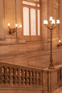 An elegant, historic interior featuring a grand staircase with ornate stone railings. The space is illuminated by intricately designed, vintage wall lamps and floor lamps with warm, glowing bulbs. Large windows with decorative curtains add to the opulent atmosphere.