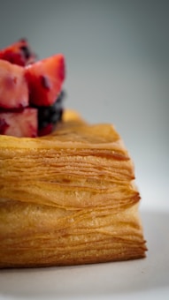 A golden-brown, flaky pastry topped with a few pieces of diced strawberries and blackberries. The focus is on the texture of the pastry layers, creating a warm and inviting feel.