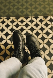 A pair of black leather boots, worn by someone wearing light-colored jeans, are positioned on a patterned tile floor featuring a repetitive circular design with black and white colors.