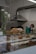 A bakery counter displays a variety of freshly baked pastries, including croissants and loaves of bread. In the background, there are industrial kitchen elements like a metal ventilation hood. A vase with white flowers and catkins decorates the counter, adding a touch of elegance.