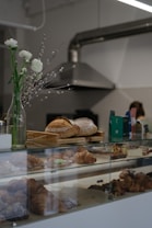 A bakery counter displays a variety of freshly baked pastries, including croissants and loaves of bread. In the background, there are industrial kitchen elements like a metal ventilation hood. A vase with white flowers and catkins decorates the counter, adding a touch of elegance.