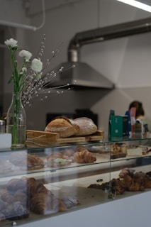 A bakery counter displays a variety of freshly baked pastries, including croissants and loaves of bread. In the background, there are industrial kitchen elements like a metal ventilation hood. A vase with white flowers and catkins decorates the counter, adding a touch of elegance.
