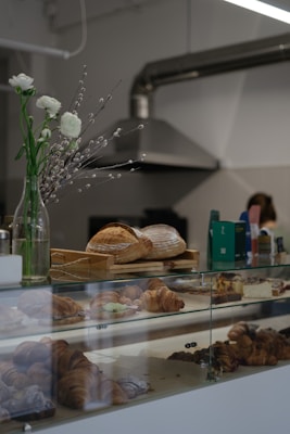 A bakery counter displays a variety of freshly baked pastries, including croissants and loaves of bread. In the background, there are industrial kitchen elements like a metal ventilation hood. A vase with white flowers and catkins decorates the counter, adding a touch of elegance.