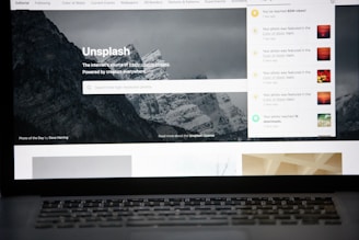 A laptop screen displays the homepage of the Unsplash website. The site features a search bar over an image of a snowy mountain landscape with trees. Notifications appear on the right side of the screen indicating user achievements and updates.