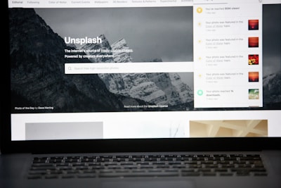 A laptop screen displays the homepage of the Unsplash website. The site features a search bar over an image of a snowy mountain landscape with trees. Notifications appear on the right side of the screen indicating user achievements and updates.