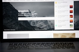 A laptop screen displays the homepage of the Unsplash website. The site features a search bar over an image of a snowy mountain landscape with trees. Notifications appear on the right side of the screen indicating user achievements and updates.