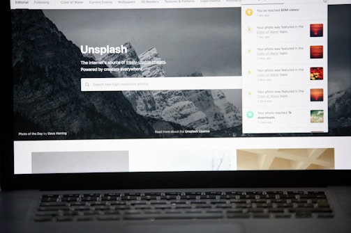A laptop screen displays the homepage of the Unsplash website. The site features a search bar over an image of a snowy mountain landscape with trees. Notifications appear on the right side of the screen indicating user achievements and updates.