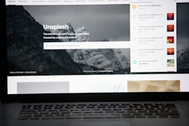 A laptop screen displays the homepage of the Unsplash website. The site features a search bar over an image of a snowy mountain landscape with trees. Notifications appear on the right side of the screen indicating user achievements and updates.