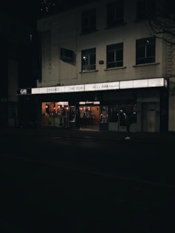 A dimly lit street scene featuring the facade of a cinema. The marquee displays movie titles such as 'Cyrano', 'The Duke', and 'Ali & Ava Preview'. The building is modest with several windows and the entrance is illuminated. A person is visible in front of the cinema.