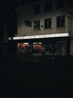A dimly lit street scene featuring the facade of a cinema. The marquee displays movie titles such as 'Cyrano', 'The Duke', and 'Ali & Ava Preview'. The building is modest with several windows and the entrance is illuminated. A person is visible in front of the cinema.