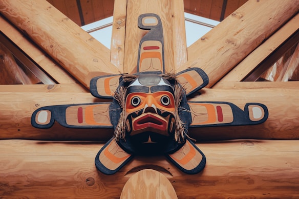 An intricately designed indigenous mask with a sun motif, featuring bold geometric patterns in shades of red, black, and orange, mounted against wooden beams.