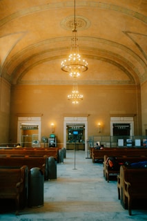 A spacious and elegant interior of a historical train station features grand chandeliers hanging from a high, ornate ceiling. The walls are adorned with warm tones, and rows of wooden benches are neatly aligned, suggesting a waiting area. At the back, there are signs for 'Tickets & Baggage' above counters, indicating a service area.