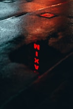 A wet street with a dark surface reflecting the red neon letters spelling 'TAXI'. The reflection is bright against the dark pavement, and the image conveys an urban atmosphere.