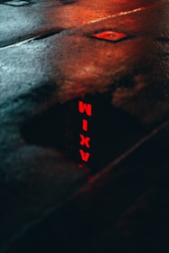 A wet street with a dark surface reflecting the red neon letters spelling 'TAXI'. The reflection is bright against the dark pavement, and the image conveys an urban atmosphere.
