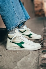 A close-up of someone wearing high-top sneakers with a white base and green accents on the side. The shoes have white laces and the person's jeans are wide-legged and reach just above the sneakers. The background is an outdoor urban setting with a stone or concrete surface.