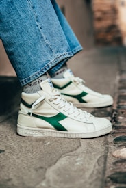 A close-up of someone wearing high-top sneakers with a white base and green accents on the side. The shoes have white laces and the person's jeans are wide-legged and reach just above the sneakers. The background is an outdoor urban setting with a stone or concrete surface.
