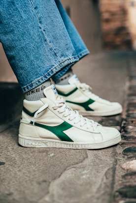 A close-up of someone wearing high-top sneakers with a white base and green accents on the side. The shoes have white laces and the person's jeans are wide-legged and reach just above the sneakers. The background is an outdoor urban setting with a stone or concrete surface.