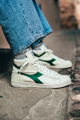 A close-up of someone wearing high-top sneakers with a white base and green accents on the side. The shoes have white laces and the person's jeans are wide-legged and reach just above the sneakers. The background is an outdoor urban setting with a stone or concrete surface.