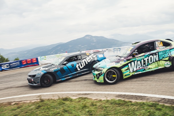 Two racing cars are seen speeding on a track, surrounded by mountainous scenery. The cars are painted with bold logos and designs, suggesting sponsorship in a motor sport event. A guardrail and various banners can be seen along the track.