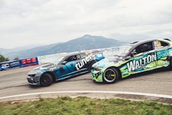 Two racing cars are seen speeding on a track, surrounded by mountainous scenery. The cars are painted with bold logos and designs, suggesting sponsorship in a motor sport event. A guardrail and various banners can be seen along the track.
