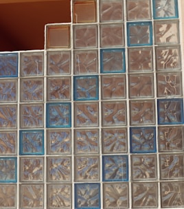 A wall constructed of glass blocks arranged in a grid pattern, featuring a mix of clear and blue-tinted blocks. The texture and reflections within the blocks create a shimmering and distorted view, contributing to an abstract visual effect.