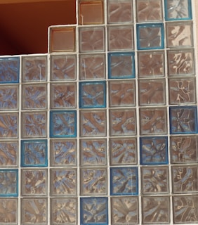 A wall constructed of glass blocks arranged in a grid pattern, featuring a mix of clear and blue-tinted blocks. The texture and reflections within the blocks create a shimmering and distorted view, contributing to an abstract visual effect.