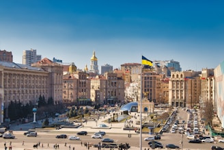 A busy urban square with a large flagpole flying the Ukrainian flag. In the center, there is a glass dome structure surrounded by people and parked cars. The background features historic buildings with ornate architectural details, and a gold-domed building is prominently visible. The sky is clear and blue, suggesting a sunny day.