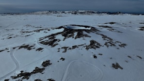 A vast snowy landscape featuring a prominent crater-like formation in the center. The scene is mostly barren with scattered dark patches of rock, suggesting a desolate and remote environment. The horizon shows distant mountain ranges under a cloudy sky.