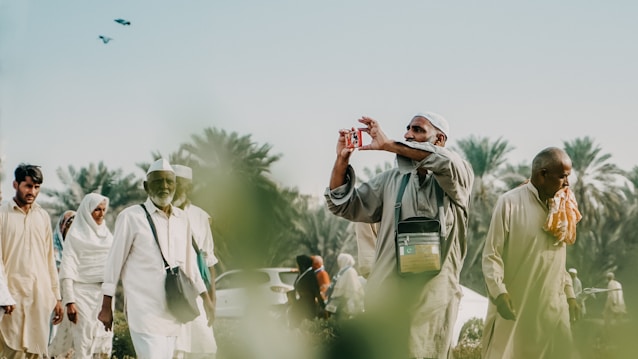 A group of people, mostly men, walk in an outdoor setting with palm trees in the background. One man is taking a photo with a small camera, while others appear to be conversing and walking. They are dressed in light-colored traditional clothing, and there are birds flying in the sky above.