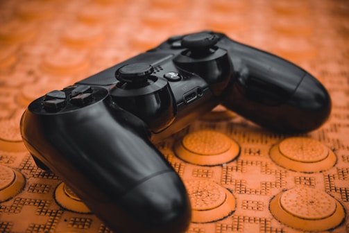 A black video game controller lies on a textured, orange surface with circular patterns. The controller features prominent buttons and analog sticks, appearing slightly glossy against the contrasting background.