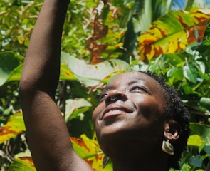 An individual with short hair and a nose ring is smiling with their face turned upwards, surrounded by lush green foliage in a tropical setting.