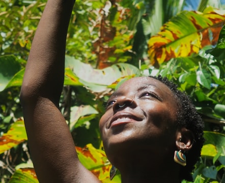 An individual with short hair and a nose ring is smiling with their face turned upwards, surrounded by lush green foliage in a tropical setting.