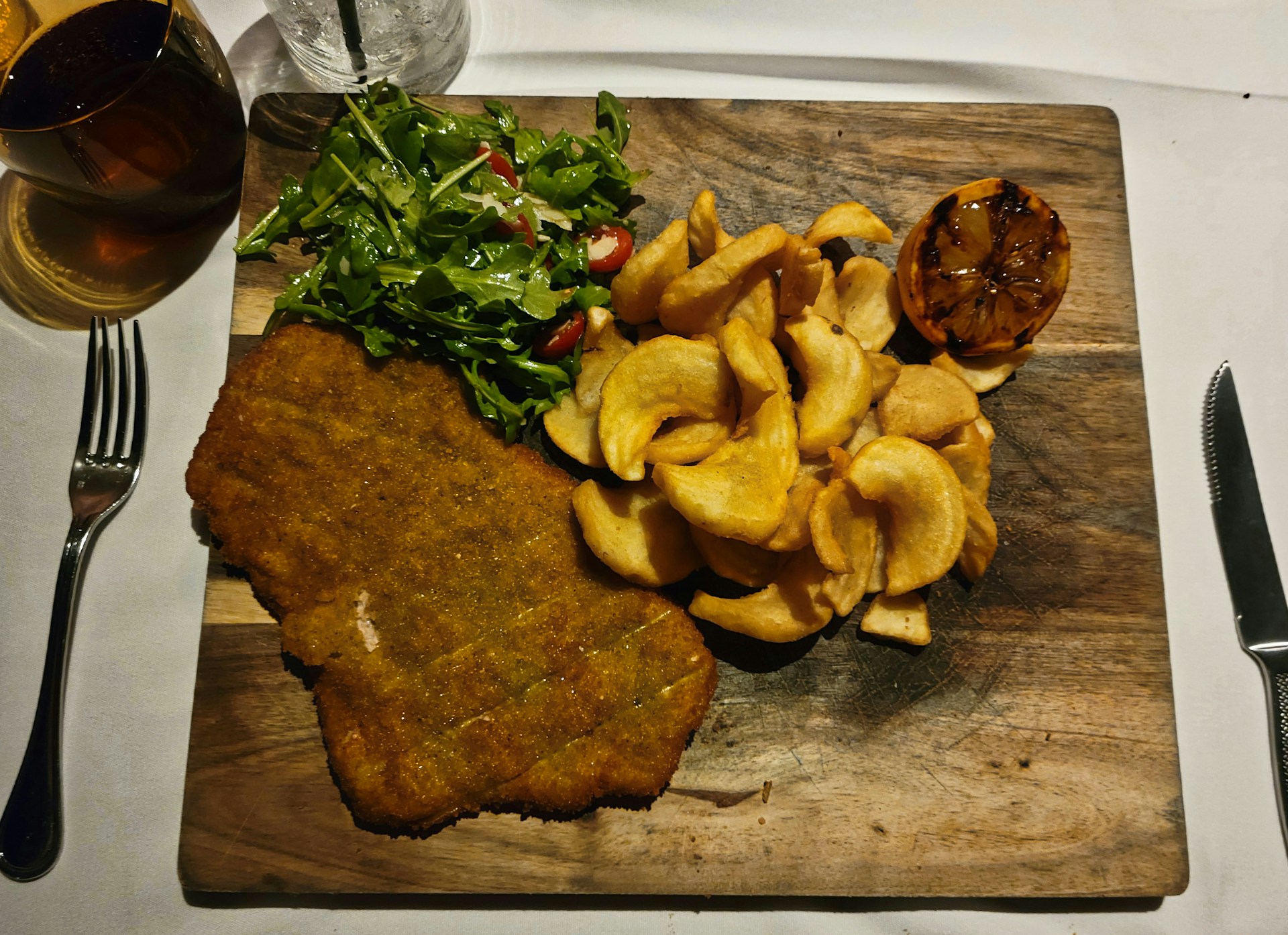 A close-up of a golden, crispy milanesa served with a fresh wedge of lemon and a side of rustic fries on a wooden table.