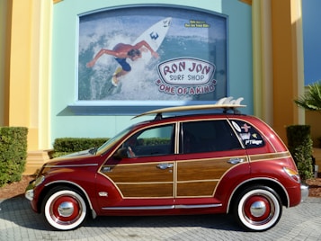 A custom red car with wood paneling is parked in front of a colorful building. The car has a surfboard mounted on its roof and various stickers on the rear window. Behind it, a large sign for 'Ron Jon Surf Shop' is displayed, featuring an image of a surfer riding a wave.