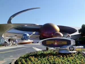A futuristic attraction featuring large, spherical structures with a metallic appearance, resembling planets with rings. A sign in the foreground reads 'Mission: SPACE' with a logo. Visitors walk towards the entrance under a sunny sky.