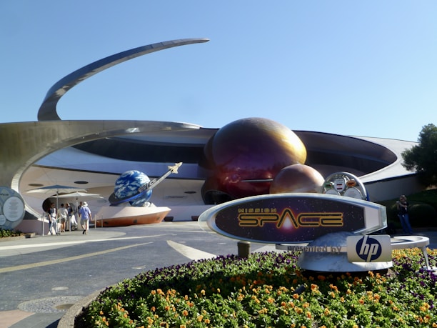 A futuristic attraction featuring large, spherical structures with a metallic appearance, resembling planets with rings. A sign in the foreground reads 'Mission: SPACE' with a logo. Visitors walk towards the entrance under a sunny sky.